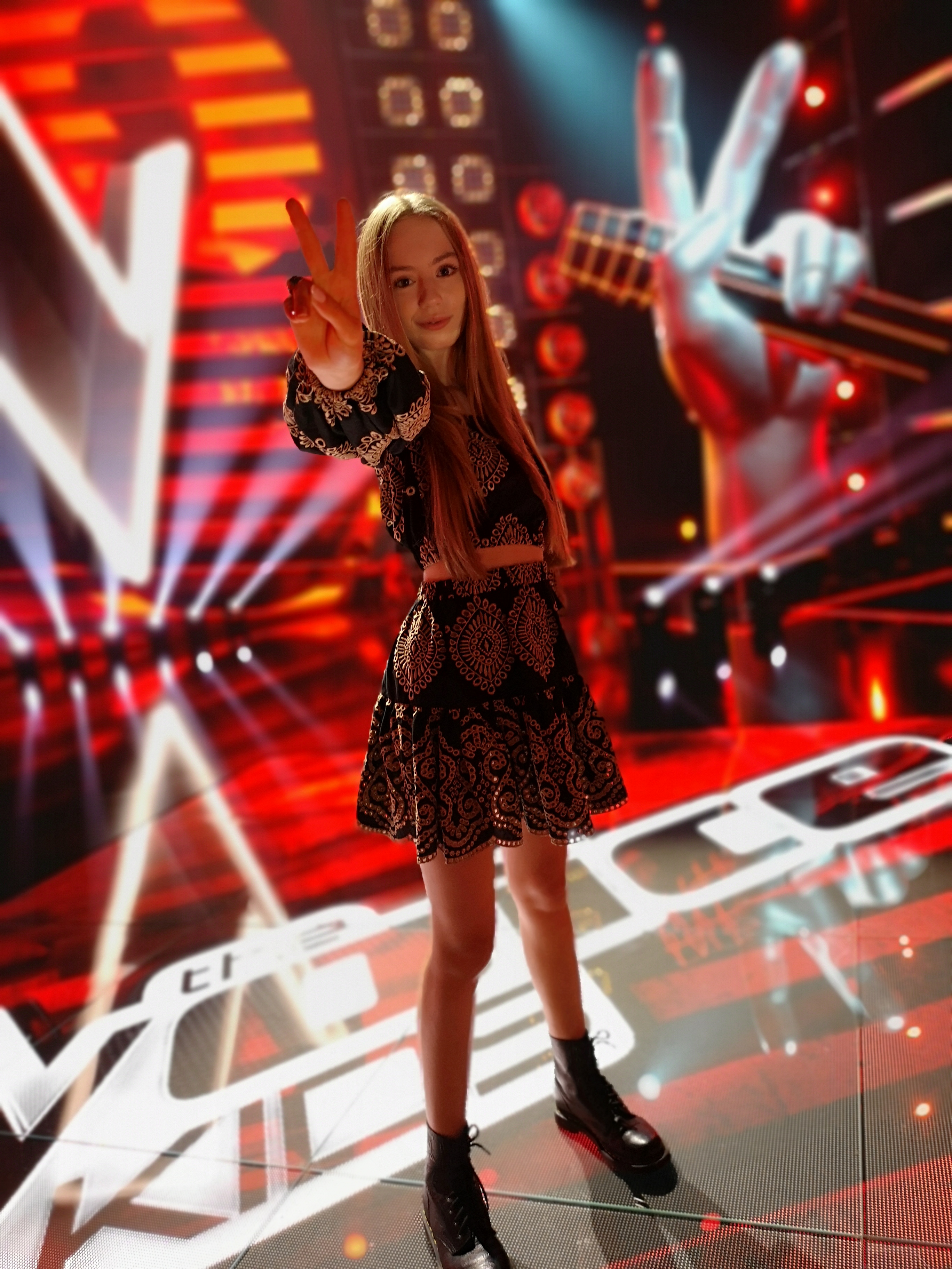 Hania Sztachańska na scenie programu The Voice Kids i pokazująca literę "V". W tle widoczny symbol programu - palce dłoni trzymającej mikrofon ułożone w literę "V".
