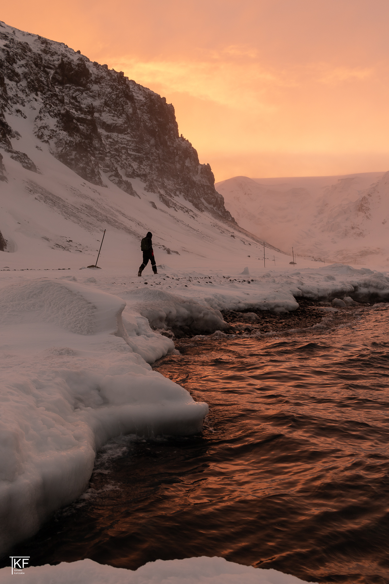 Na zdjęciu idący mężczyzna, dookoła pokrywa śnieżna, i woda, w której odbija się wieczorne, pomarańczowe światło, niebo również w kolorze pomarańczowym.