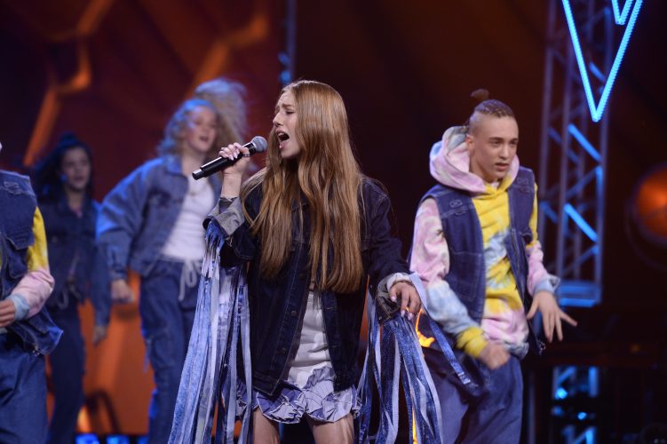 Hania Sztachańska podczas występu na scenie w programie The Voice Kids - Hania śpiewa, za nią widać dwie osoby z zespołu tanecznego w trakcie występu