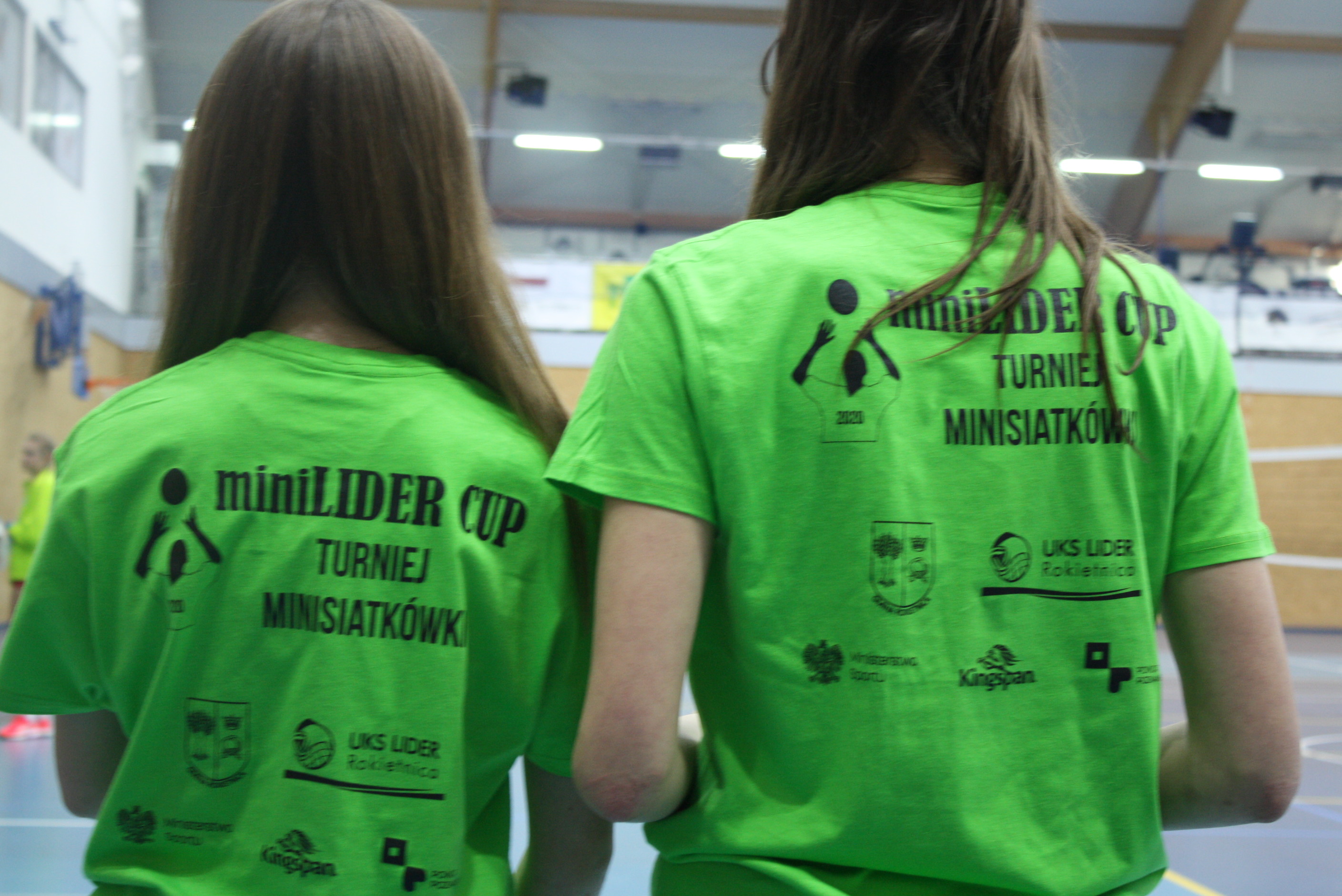 Na zdjęciu znajdują się dwie dziewczynki stojące tyłem do obiektywu. Mają na sobie zielone koszulki z napisem miniLIDER CUP TURNIEJ MINISIATKÓWKI, rysunkiem siatkarza i logotypami organizatorów i sponsorów: Gmina Rokietnica, UKS Lider, Ministerstwo Sportu, Kingspan, Powiat Poznański.