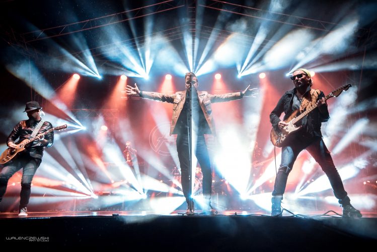 zdjęcie przedstawia zespół FEEL na scenie podczas wykonywania utworu. Tło jest czarne rozświetlone niebieskimi i czerwonymi światłami