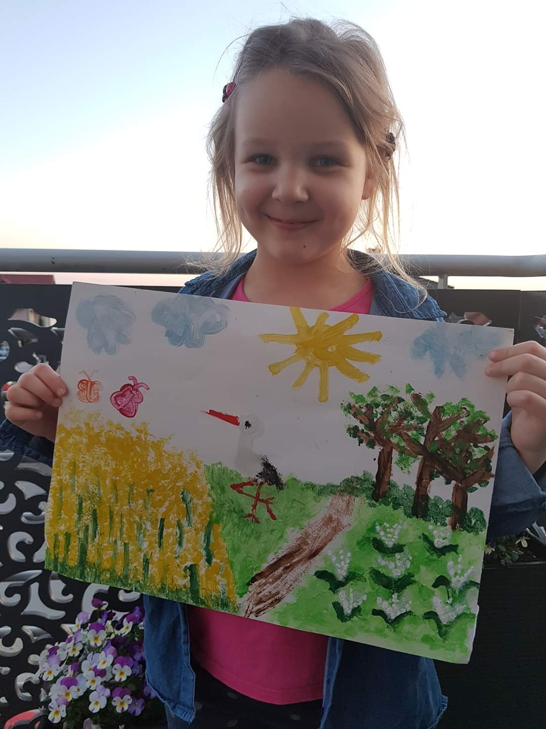 Na zdjęciu widoczna uśmiechnięta dziewczynka prezentująca namalowany przez siebie obraz. Na nim znajduje się wiosenny krajobraz - łąka, drzewa, kwiaty, bocian, rzepak, niebo, słonce, chmury i motyle.