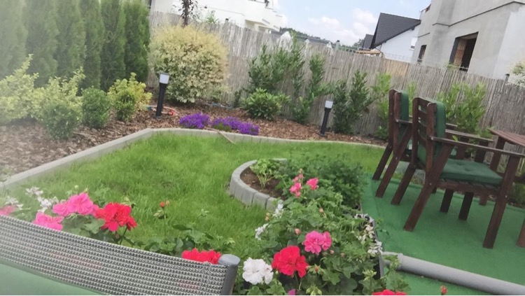 Na zdjęciu widać ogród z posadzonymi różami, ozdobnymi krzewami i trawami, po prawej stronie widać brązowe, drewniane krzesełka ogrodowe.