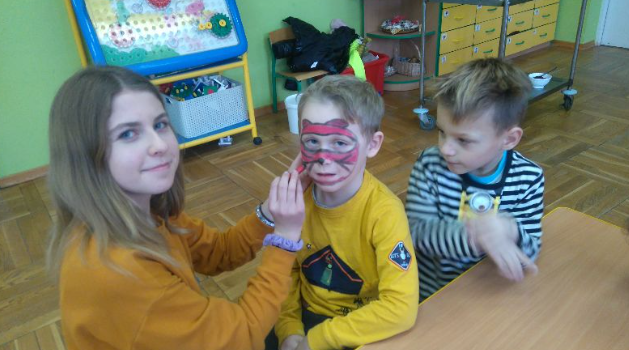Na zdjęciu młoda dziewczyna malująca twarz chłopcu w przedszkolu "Bajeczka" w Rokietnicy. Oboje patrzą w kierunku fotografa. Obok siedzi drugi chłopiec i patrzy w bok. Autor: Stowarzyszenie CREO