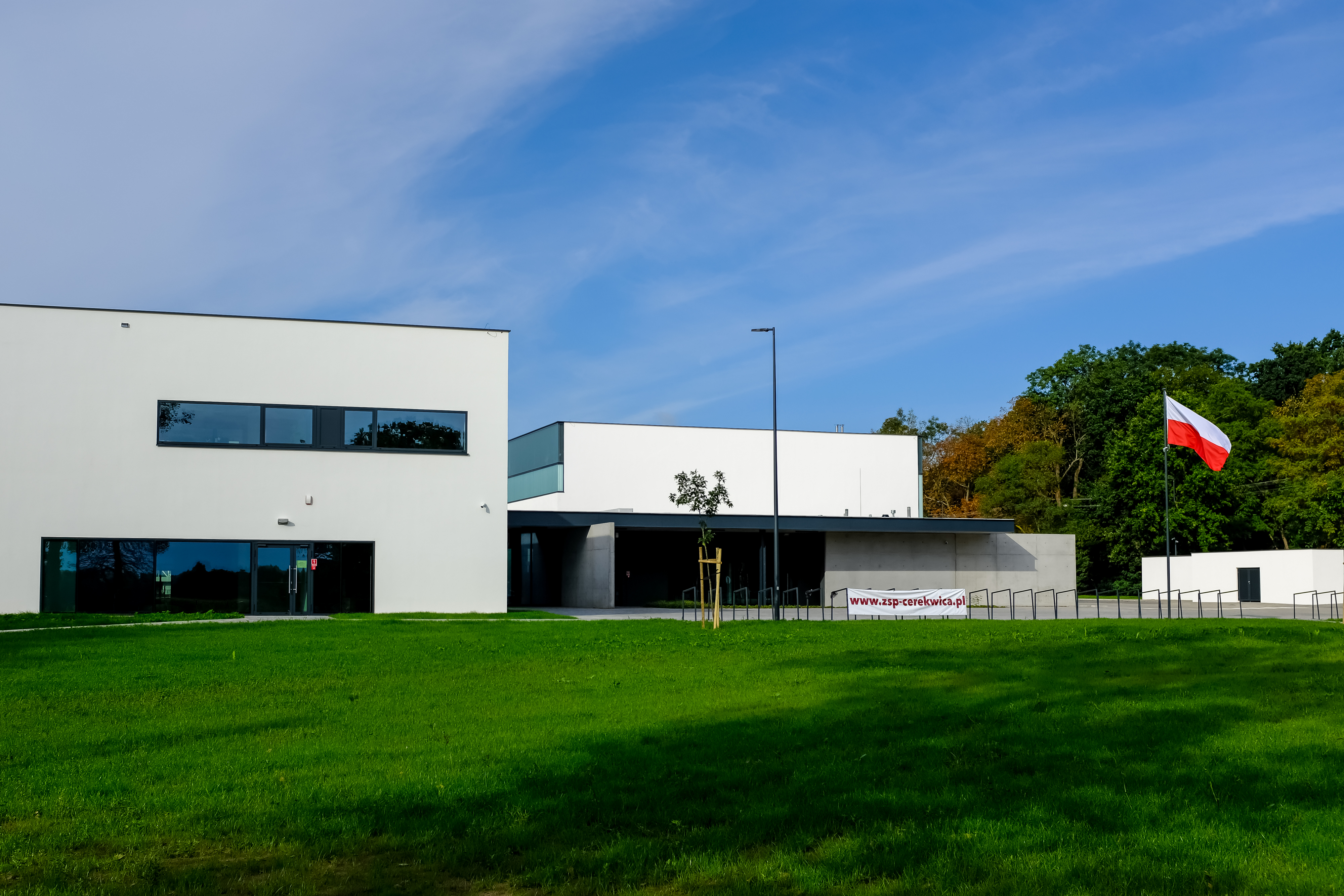 Na zdjęciu widać budynek Szkoły Podstawowej w Cerekwicy. Przed szkołą widoczny szeroki trawnik, po prawej stronie masz z wywieszoną biało-czerwoną flagą.