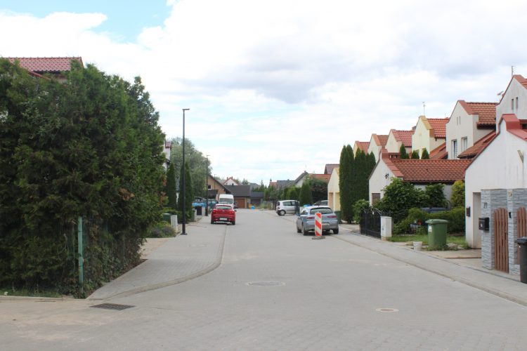 Zdjęcie przedstawia ulicę. Po obu jej stronach są chodniki, po lewej stronie żywopłot, po prawej zabudowa mieszkalna. Na poboczach stoją zaparkowane auta.