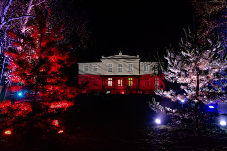Na zdjęciu widać budynek dworku dwupiętrowego nocą, podświetlonego na biało-czerwono, zgodnie z kolorystyką polskiej flagi narodowej. Po lewej stronie zdjęcia widać choinkę podświetloną na czerwono, po prawej stronie zdjęcia na pirrwszym planie widać choinkę podświetloną na biało.