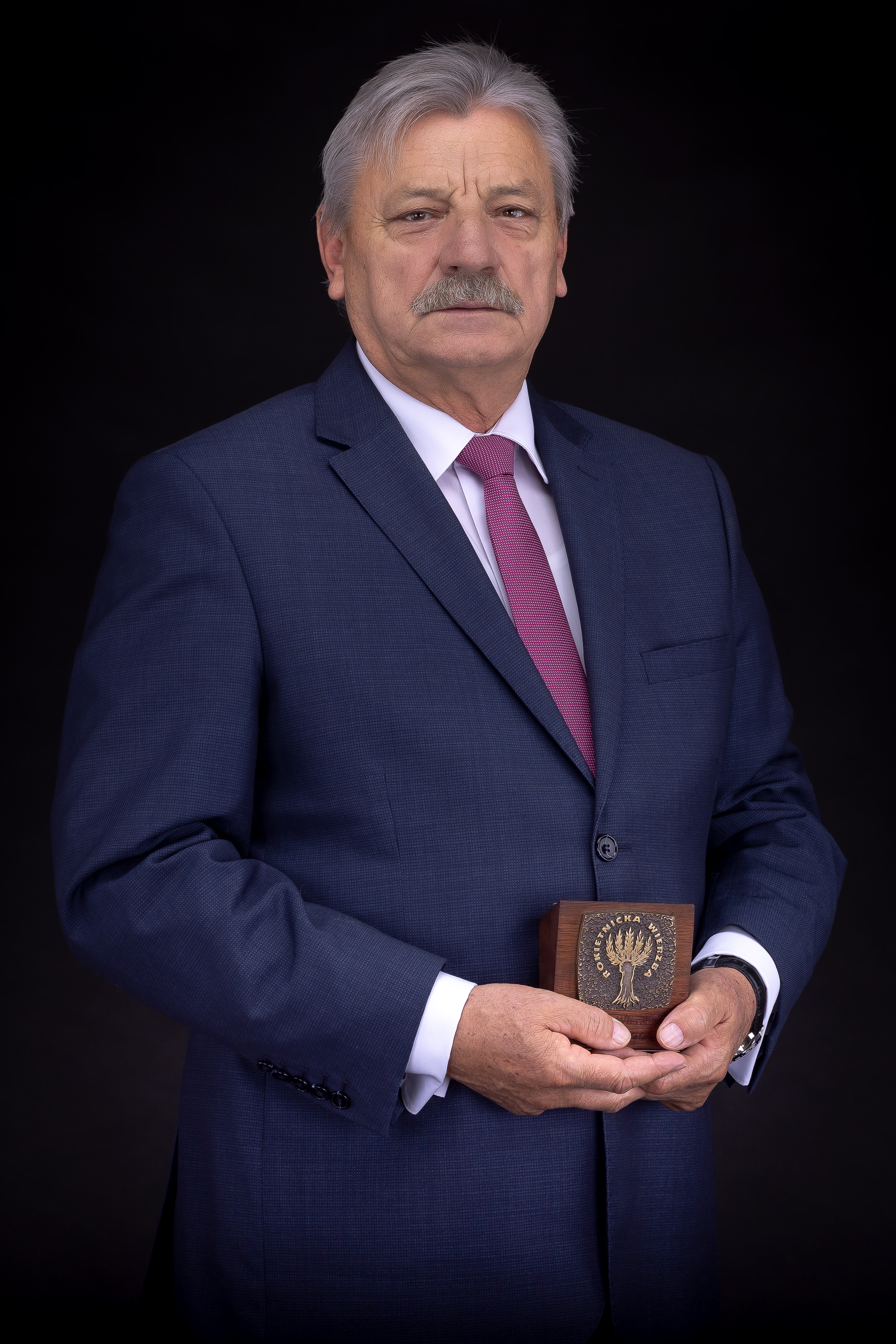 Na zdjęciu laureat statuetki Rokietnickiej Wierzby 2021, Wojciech Tulibacki, na czarnym tle w granatowym garniturze, białej koszuli i różowym krawacie. W rękach trzyma statuetkę Rokietnickiej Wierzby.
