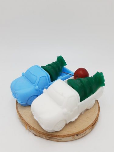 Na zdjęciu widać dwa mydełka naturalne w kształcie samochodów z choinką na pace. Jeden samochód jest biały, jeden niebieski, oba stoją na płacie drewna.