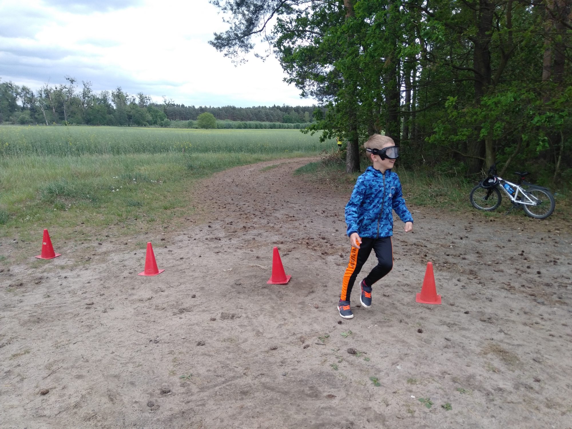 Na zdjęciu chłopiec w alkogoglach, pokonujący slalom, wyznaczony przez pachołki. Dalej rower i las. Po lewej stronie łąka i dalej las.
