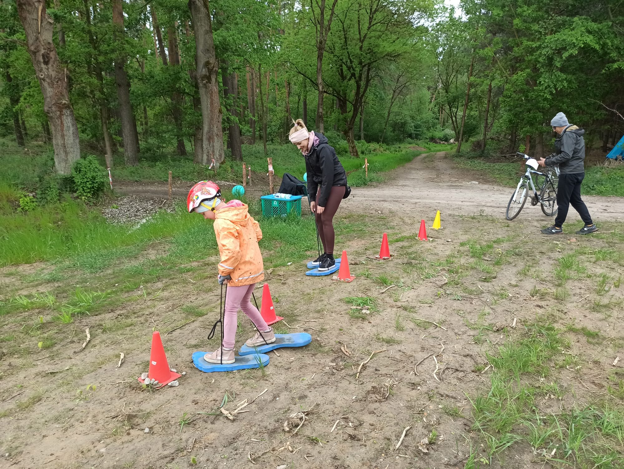 Na zdjęciu 3 osoby - kobieta, mężczyzna i dziewczynka. Kobieta i dziewczynka idą slalomem z wykorzystaniem dużych stóp, które podnoszą poprzez pociąganie za sznurki. Za nimi mężczyzna stojący przy rowerach i las.