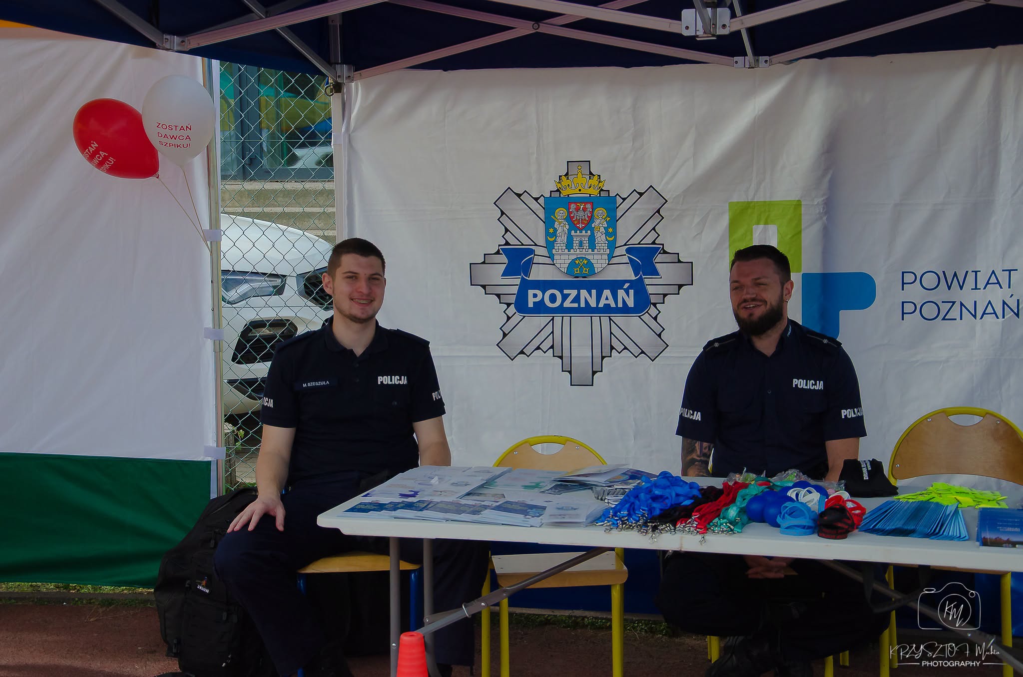 Na zdjęciu dw&oacute;ch policjant&oacute;w siedzących przy stoliku, na&nbsp;kt&oacute;rym są rozłożone gadżety. Za nimi baner z&nbsp;symbolem policji w&nbsp;Poznaniu.