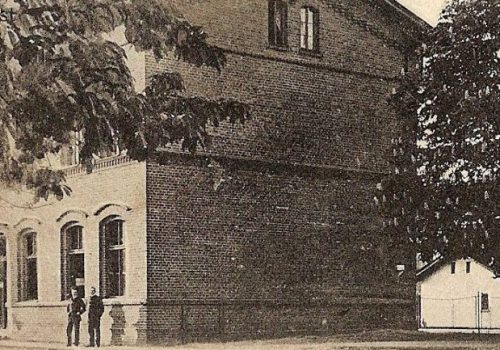 Czarno białe zdjęcie pokazujące budynek dworca w&nbsp;Rokietnicy i&nbsp;na jego tle dwie postacie