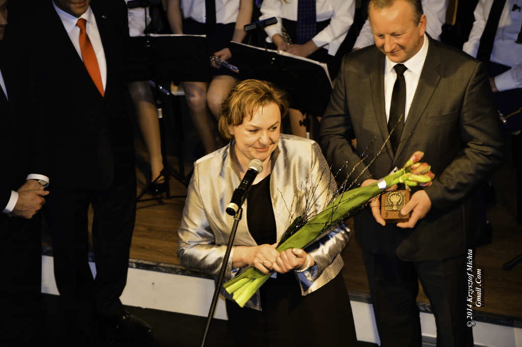 Katarzyna i Roman Gorajowie Na zdjęciu Katarzyna i Roman Gorajowie w eleganckich strojach. Mężczyzna trzyma w rękach statuetkę. Kobieta stoi przy mikrofonie na statywie. W rękach trzyma kwiaty. Za nimi muzycy na scenie.