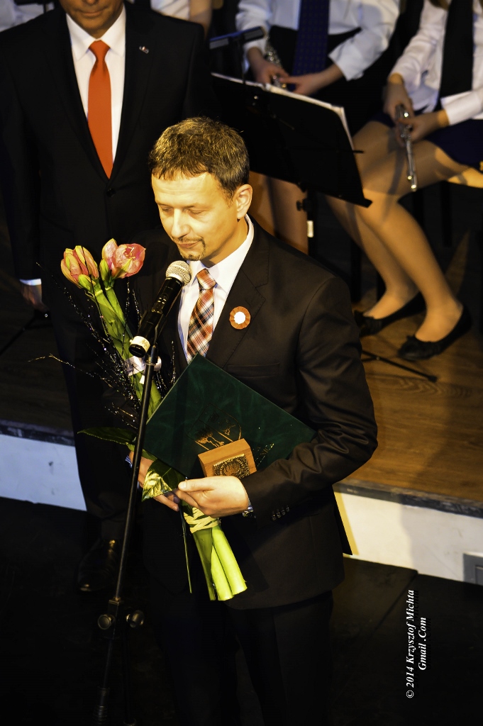 Wojciech Kaczmarek Na zdjęciu Wojciech Kaczmarek w ciemnym garniturze, białej koszuli i krawacie. W rękach trzyma teczkę, statuetkę i kwiaty. Mówi do mikrofonu na statywie. Za nim muzycy na scenie.