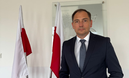 W tle herb gminy Rokietnica, na zdjęciu Wójt Gminy Rokietnica Bartosz Derech z 2 flagami Polski po lewej stronie. 
