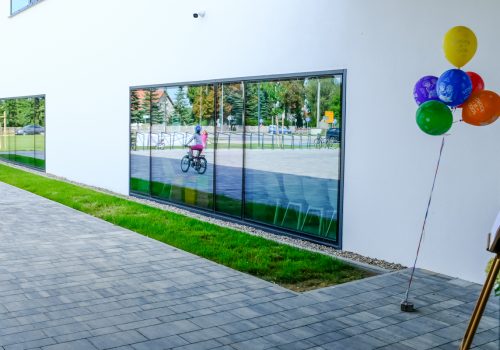 Na zdjęciu szkoła podstawowa w Cerekwicy z zewnątrz. W jednym z okien widoczne odbicie dwóch dziewczynek, z których jedna jeździ rowerem oraz stojaków na rowery. Z prawej strony okna kolorowe balony. Fot. Damian Nowicki