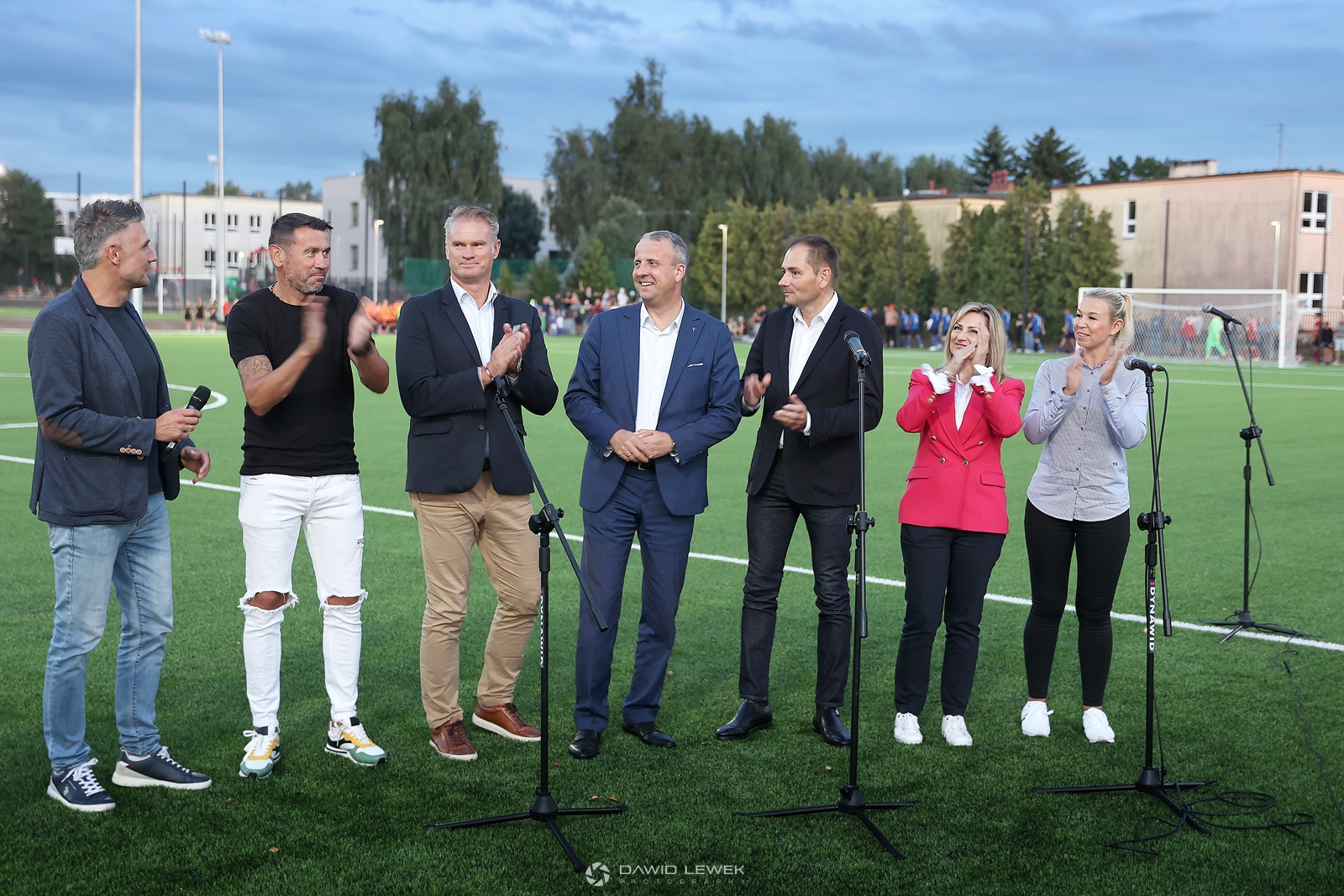 Na zdjęciu widoczna grupa ludzi stojąca na boisku do piłki nożnej - od lewej 5 mężczyzn, następnie dwie kobiety. Przed nimi cztery mikrofony na statywach.