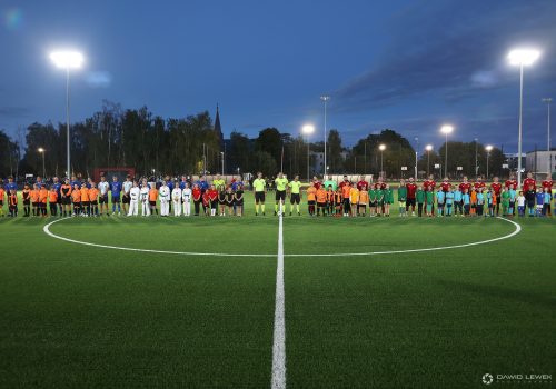 Na zdjęciu widać dwie drużyny piłkarskie stojące w rzędzie na boisku do piłki nożnej. Przed piłkarzami stoją dzieci, między drużynami stoi trzech sędziów.