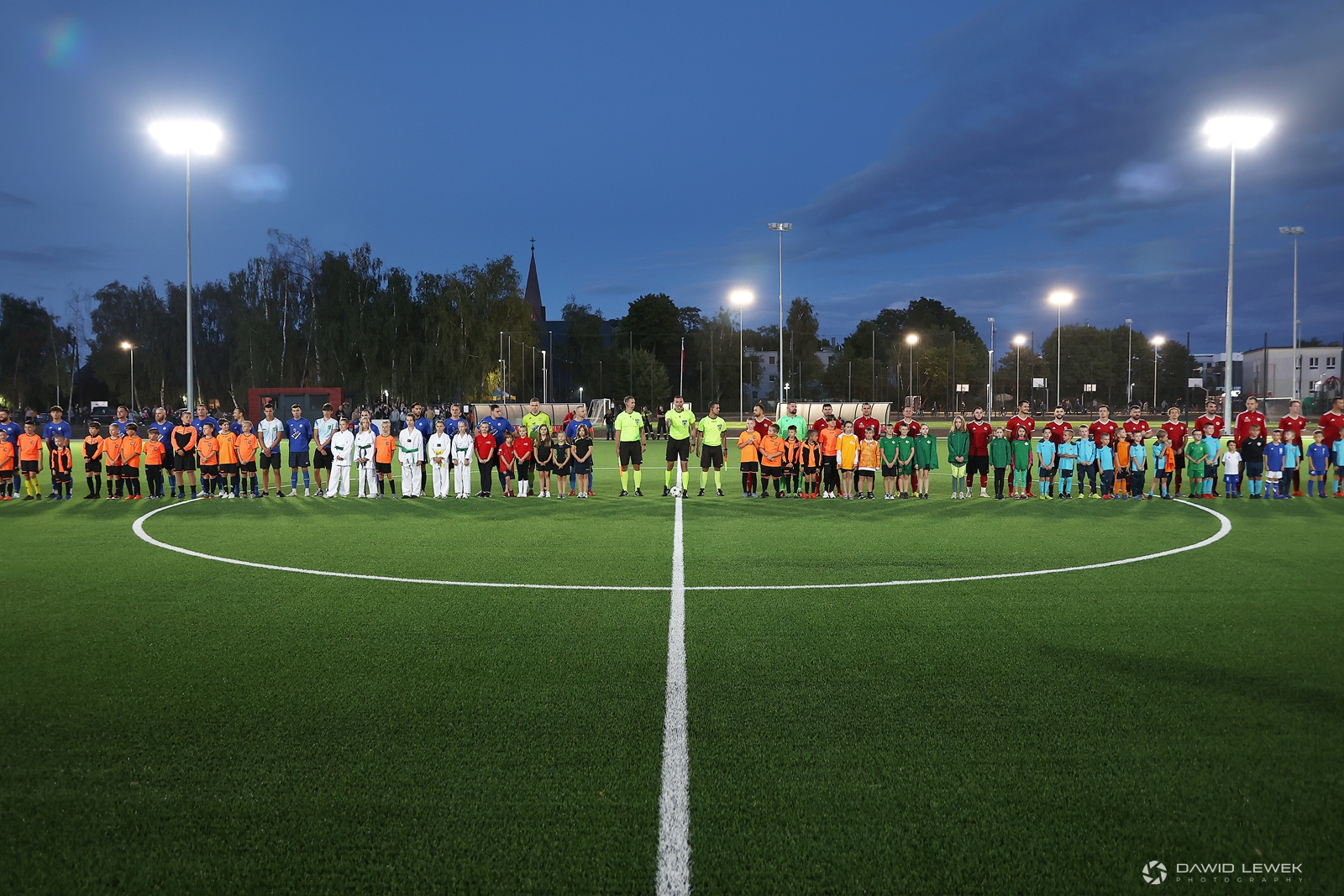 Na zdjęciu widać dwie drużyny piłkarskie stojące w rzędzie na boisku do piłki nożnej. Przed piłkarzami stoją dzieci, między drużynami stoi trzech sędziów.