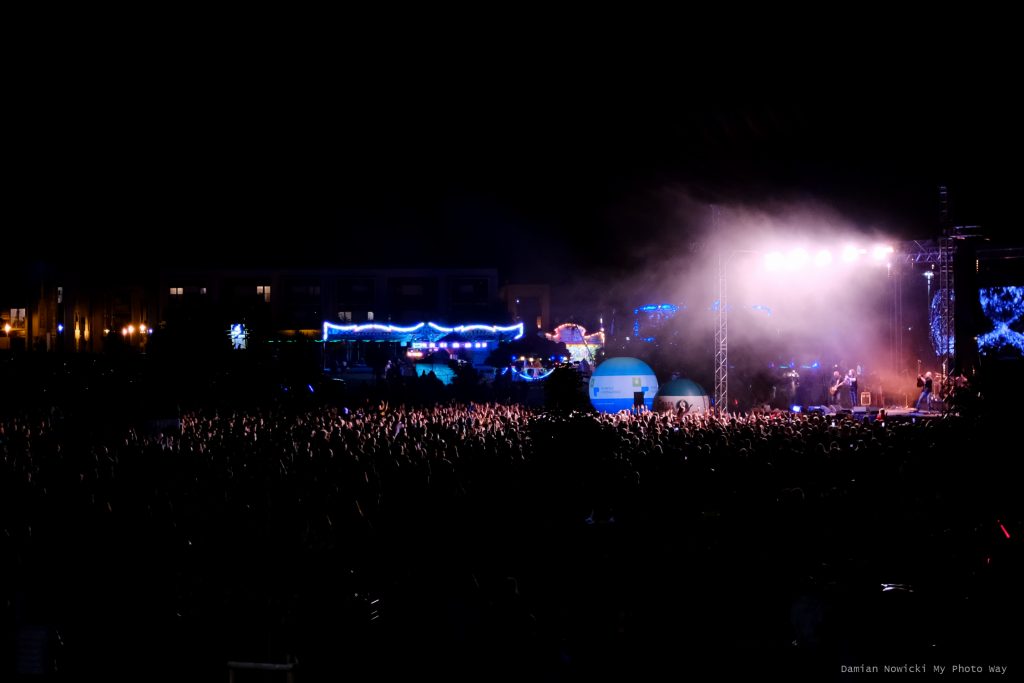 Na zdjęciu zrobionym nocą tłum podczas koncertu Piotra Cugowskiego. Na scenie zespół, przed nią balony. Dalej wesołe miasteczko.