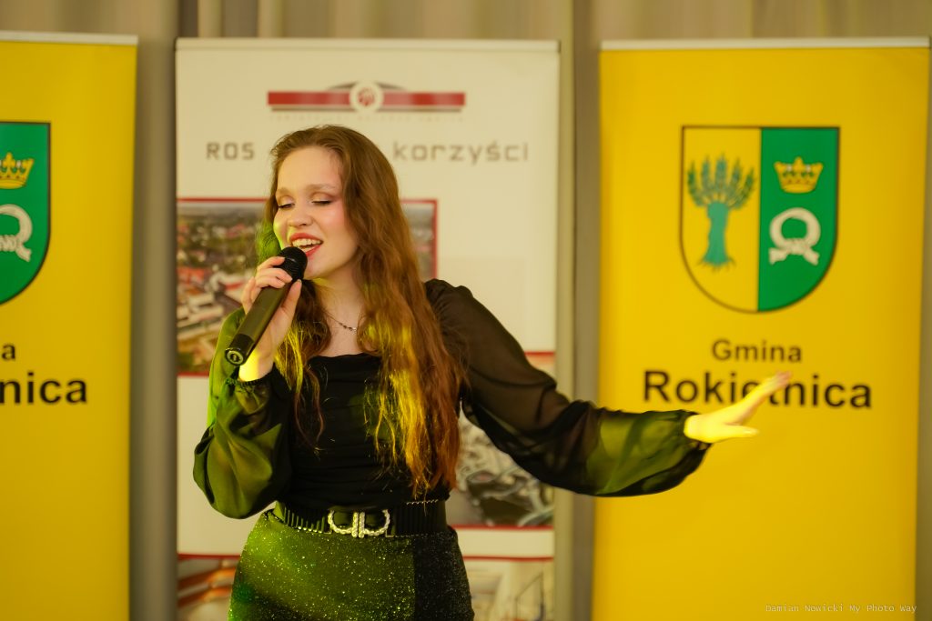 Na zdjęciu Julia Gąska śpiewająca i trzymająca mikrofon. Za nią 3 rollupy: na środku Rokietnickiego Ośrodka Sportu, po bokach Gminy Rokietnica. Fot. Damian Nowicki